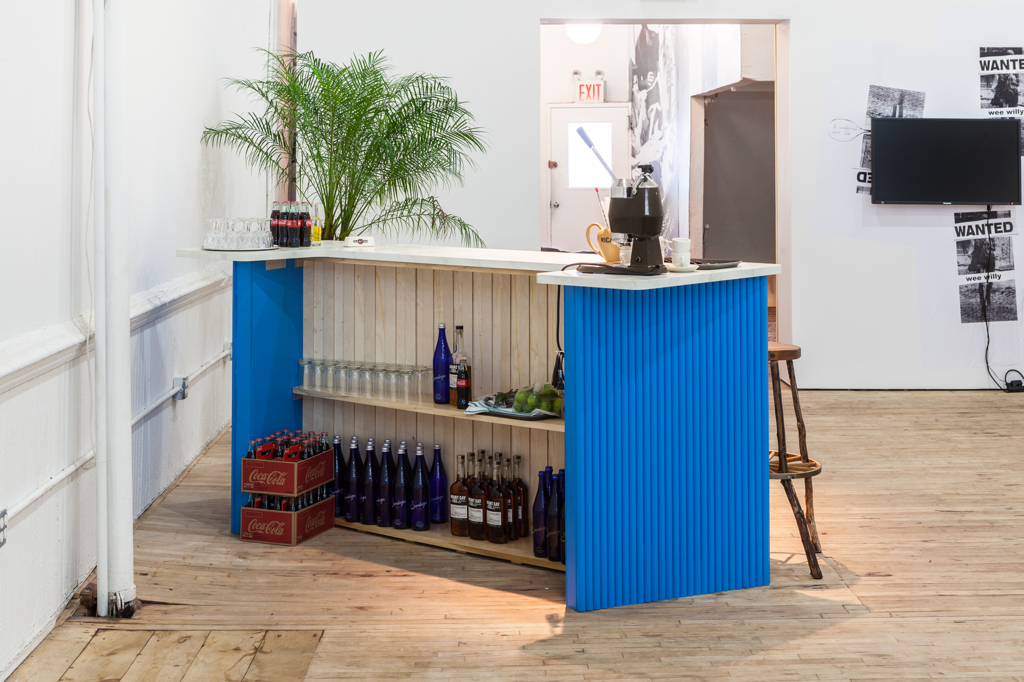 A view from behind an angled bar with blue walls. Shelves full of Coca-Cola, liquor, and glasses are visible. On the counter, more Coca-Cola bottles, glassware, and an esspresso machine. A palm leaf is positioned beyond the counter top.