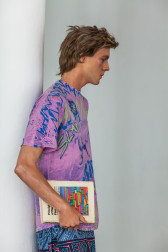A side view of a sculpture of a man leaning againt a white column. He hols a book in his right hand. He is wearing a purple shirt and blue patterned shorts.