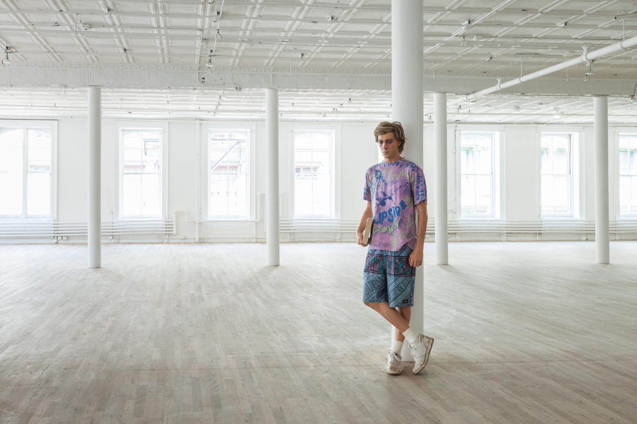 A sculpture of a man holding a book is leaning against a column in an empty, open room. He is wearing a purple shirt, patterned blue shorts, and is crossing his legs, wearing white sneakers.