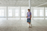 A sculpture of a man holding a book is leaning against a column in an empty, open room. He is wearing a purple shirt, patterned blue shorts, and is crossing his legs, wearing white sneakers.