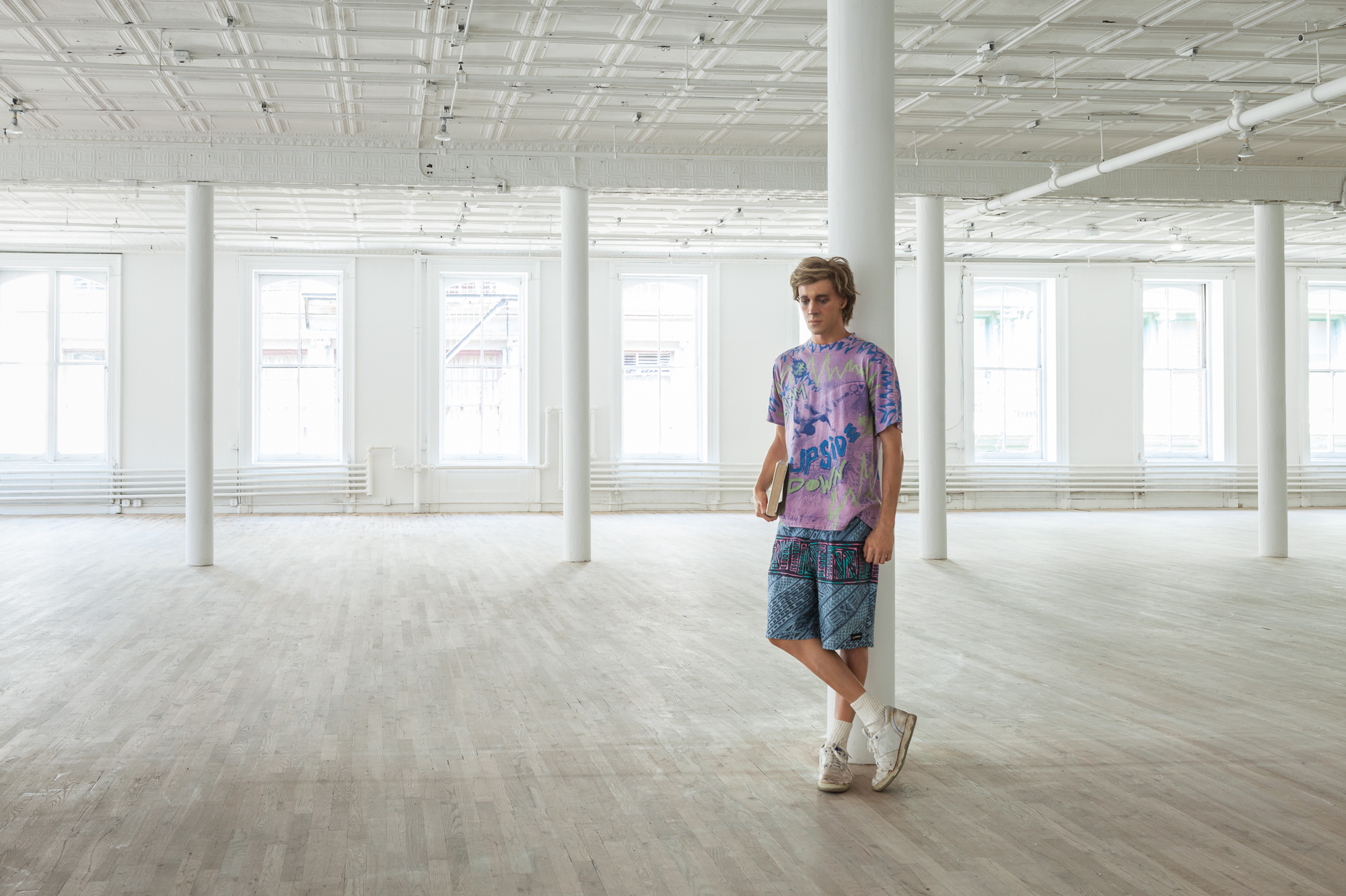 A sculpture of a man holding a book is leaning against a column in an empty, open room. He is wearing a purple shirt, patterned blue shorts, and is crossing his legs, wearing white sneakers.