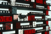 Close up image of a series of black shelves mounted to a white wall. Various wooden blocks painted with scenes of crowds, witches, and other characters are placed on the shelves, alongside smaller blocks decorated with white letters that spell out words including 