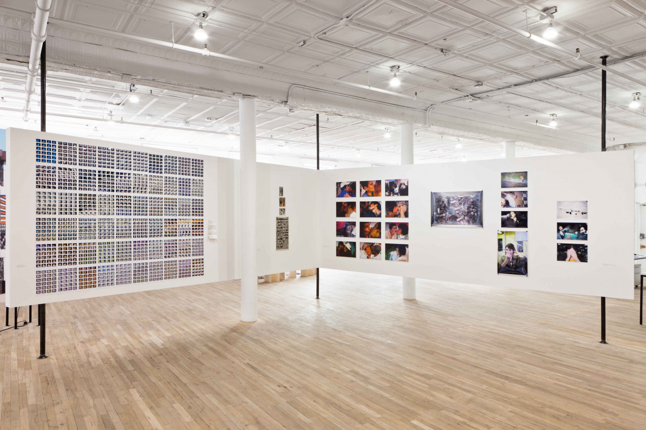 Two white walls are suspended on six vertical poles and are arranged so that they form a right corner. A large grid-like composition composed of many small squares is mounted on the left wall. On the right wall there are many photographs and printed video stills either mounted in grid patterns or one above the other.