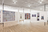 Two white walls are suspended on six vertical poles and are arranged so that they form a right corner. A large grid-like composition composed of many small squares is mounted on the left wall. On the right wall there are many photographs and printed video stills either mounted in grid patterns or one above the other.