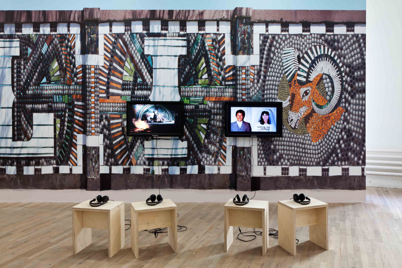 Two video monitors are mounted on a colorful gallery wall covered with a photo decal of large, white letters and a ram