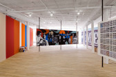 Four gallery walls sit at various angles in a bright-lit exhibition space. One of the walls is decorated with colorful vertical stripes. Another wall is covered in many small, square grid-like compositions. A wall at the photograph's center is decorated with a composition of abstract, smooth shapes and various figures rendered in black and white.