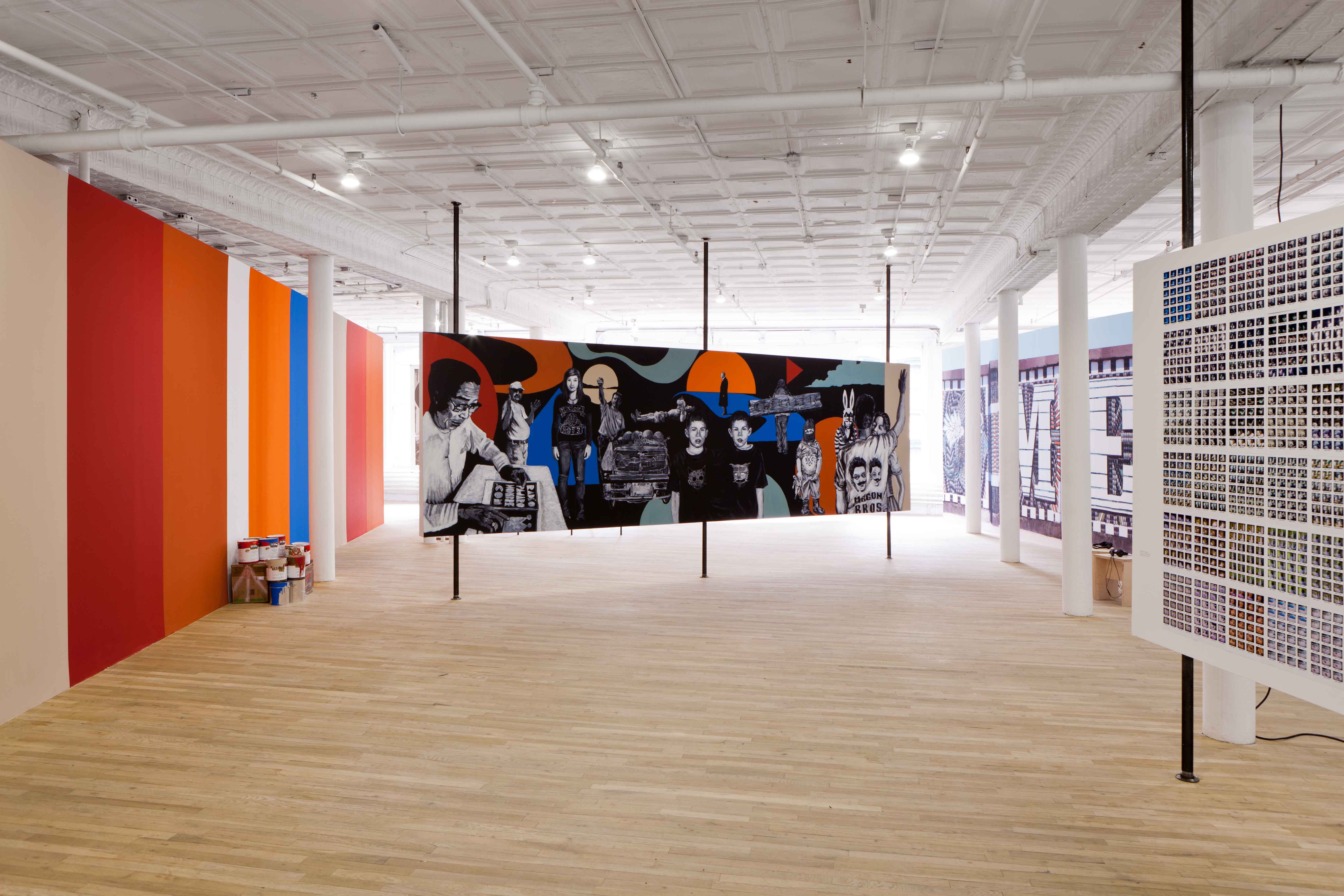 Four gallery walls sit at various angles in a bright-lit exhibition space. One of the walls is decorated with colorful vertical stripes. Another wall is covered in many small, square grid-like compositions. A wall at the photograph's center is decorated with a composition of abstract, smooth shapes and various figures rendered in black and white.