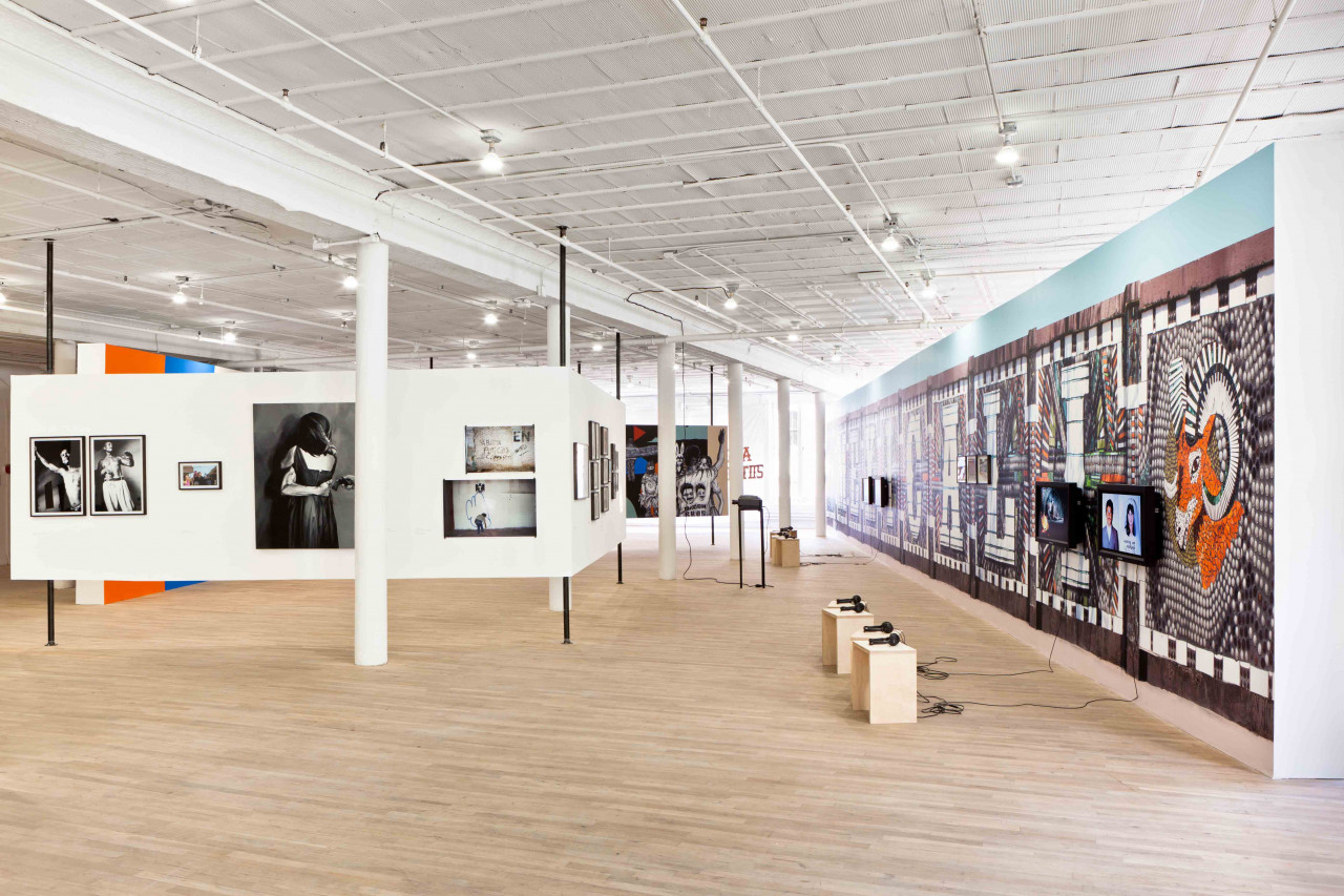 In a large, open gallery space, several photographs hang on walls to the right. To the left, multiple small monitors mounted to a colorful gallery wall play video works. In front of the monitors, there are wooden stools with headphones resting upon them.