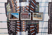 Three photographs are mounted next to one another on a colorful gallery wall, which is covered with a photo decal of a mosaic. Each of the photographs show different urban walls covered in grafitti. Graffiti on a city wall shown in the far right photograph reads, 