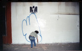 A man spray paints a large piece of abstract blue graffiti against a white city wall below a graphic emblem of an eagle. The street is dark, but he is illuminated by the flash of the camera.