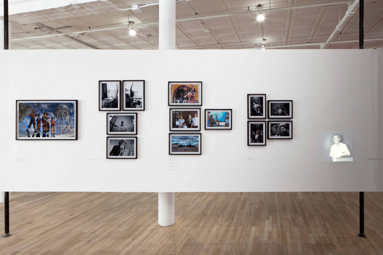 Four clusters of photographs are mounted on a white gallery wall. Some of the photographs are black and white, while others are in color. All show images of a single or groups of figures. On the far right, a small projection of a man in a white shirt illuminates the wall.