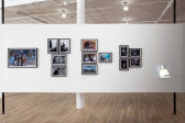 Four clusters of photographs are mounted on a white gallery wall. Some of the photographs are black and white, while others are in color. All show images of a single or groups of figures. On the far right, a small projection of a man in a white shirt illuminates the wall.