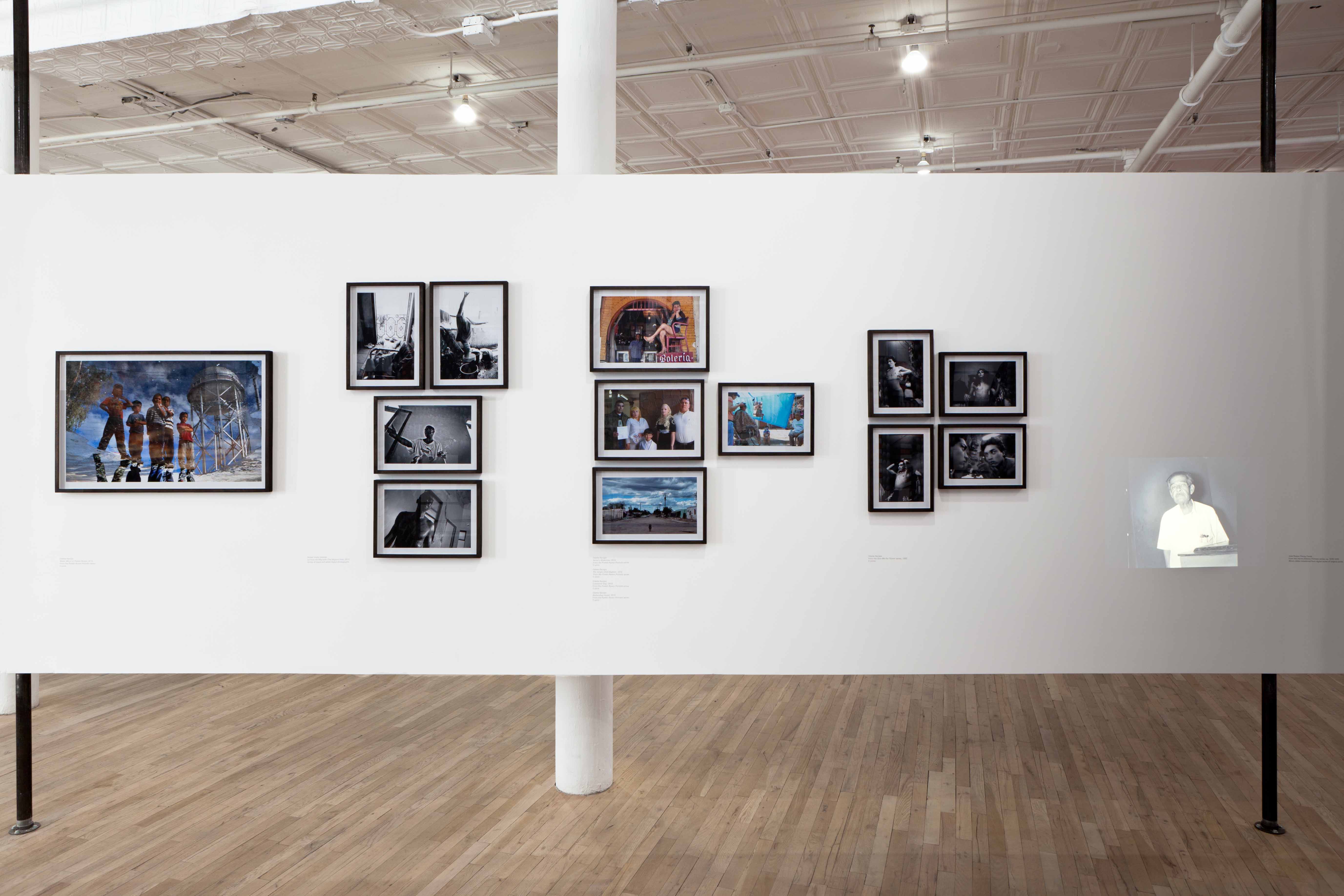 Four clusters of photographs are mounted on a white gallery wall. Some of the photographs are black and white, while others are in color. All show images of a single or groups of figures. On the far right, a small projection of a man in a white shirt illuminates the wall.