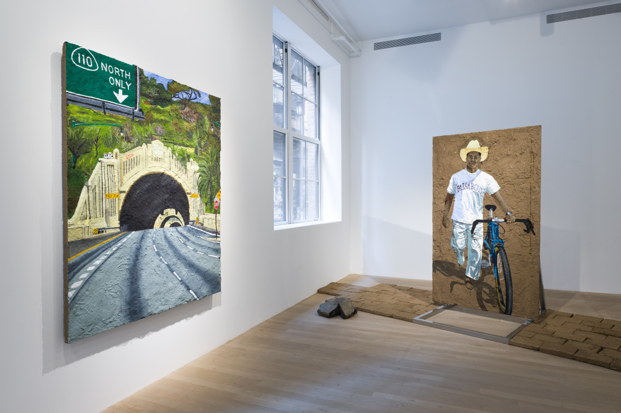 Color image of the corner of a gallery space. On the right side is a large adobe panel that rests on a steel armature. Painted onto the adobe is a portrait of a figure with a bicycle, and around the armature are adobe bricks laid out on the floor. On the lefthand wall is another adobe panel painting depicting the entrance into a freeway tunnel.