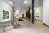 Color image of a gallery space with large panels of adobe with paintings of various figures on them. Adobe bricks are laid out on the floor to create a small walkway. Paintings are also hung on either side of the wall, each depicting different views of a freeway.