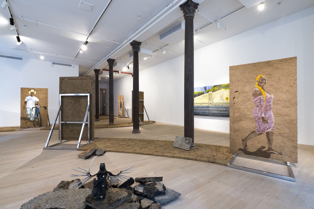 Color image of a gallery space with large panels of adobe with paintings of various figures on them. Adobe bricks are laid out on the floor to create a small walkway.