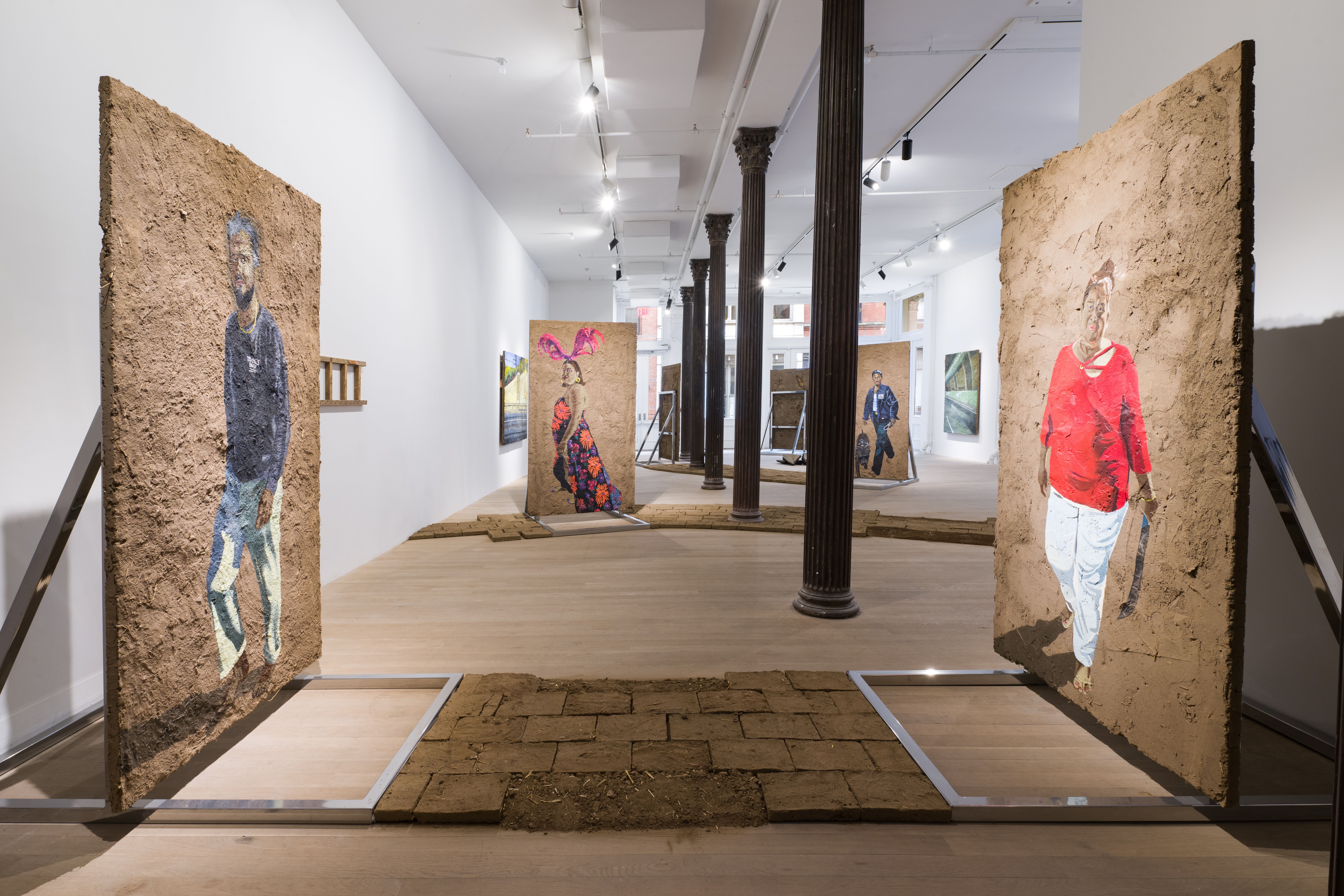 Color image of the entrance into a gallery space, with columns running down the hallway. At the forefront of the image are two large paintings made of adobe, and resting on steel armatures. Each painting features a representation of a person walking slowly. In between the armatures are adobe bricks layed out into a walkway on the floor.