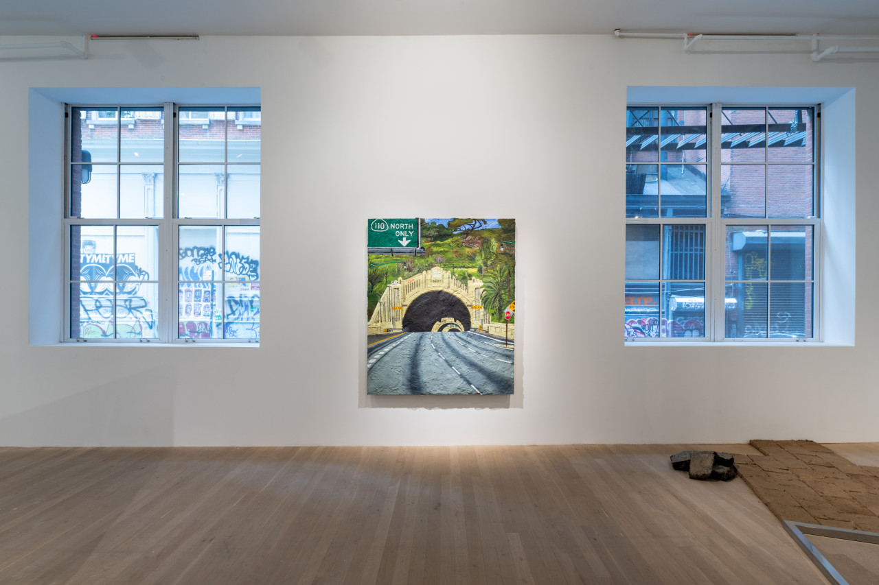 Color image of a wall of a gallery space. Hanging in the center of the wall is an adobe panel painting of the entrance to a freeway tunnel.