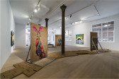 Color image of a gallery space with large paintings made on adobe panels and resting on steel armatures that are organized in various configurations. Some paintings are also hung on the wall, and adobe bricks are laid out to create a walkway in the middle of the room.