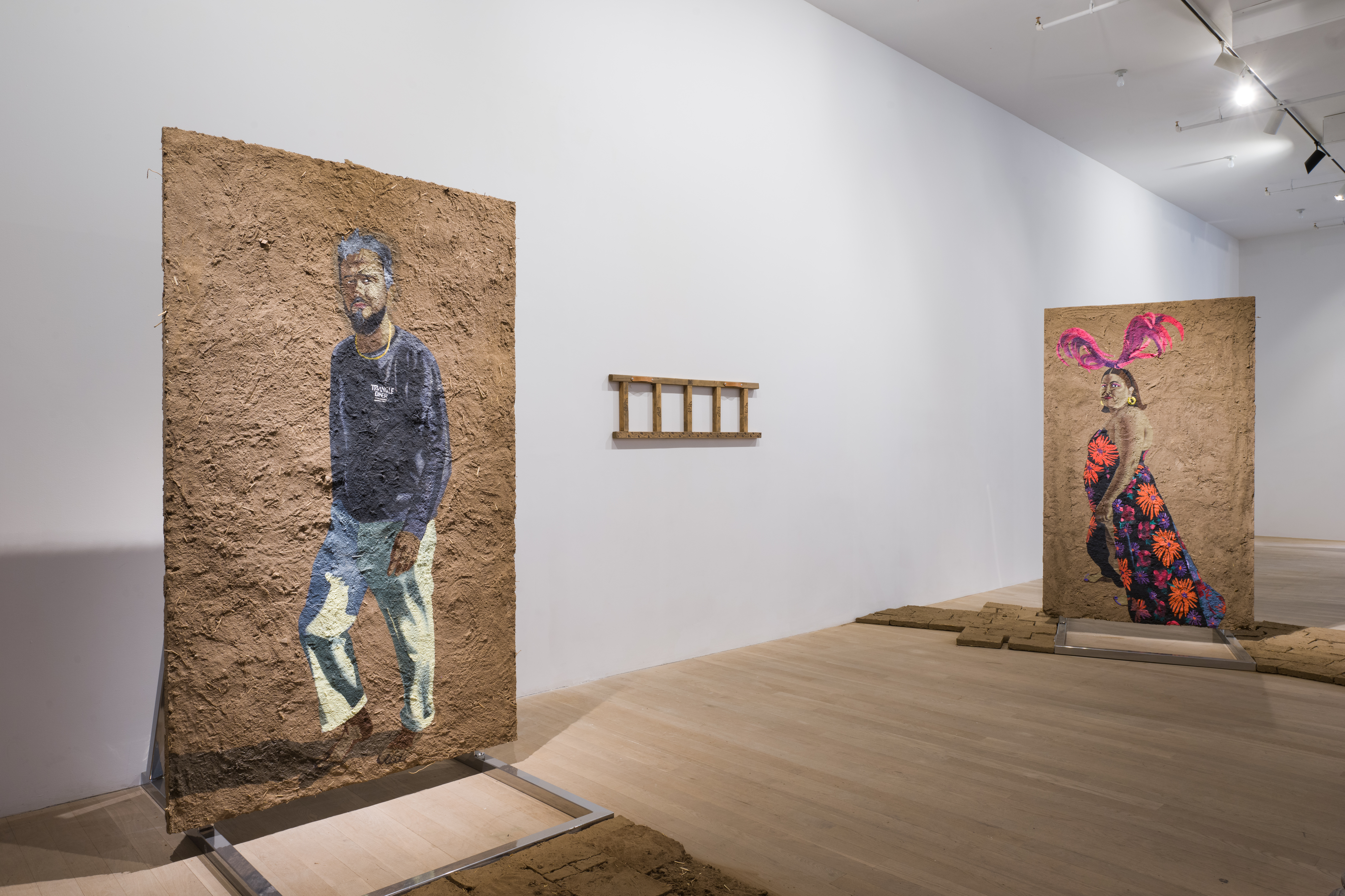 Color image of a gallery space with paintings done on adobe panels that rest on steel armatures, and are laid out around the space. The artworks depict paintings of various figures. On the wall hangs a wooden ladder horizontally.