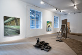 Color image of a gallery space. Along the lefthand wall are two adobe panel paintings, each depicting different parts of a freeway. In the middle of the space is a pile of freeway rubble on which sits a sculpture of a bird. Along the right side of the room are adobe bricks laid out on the floor, along with steel armatures that hold adobe panels.