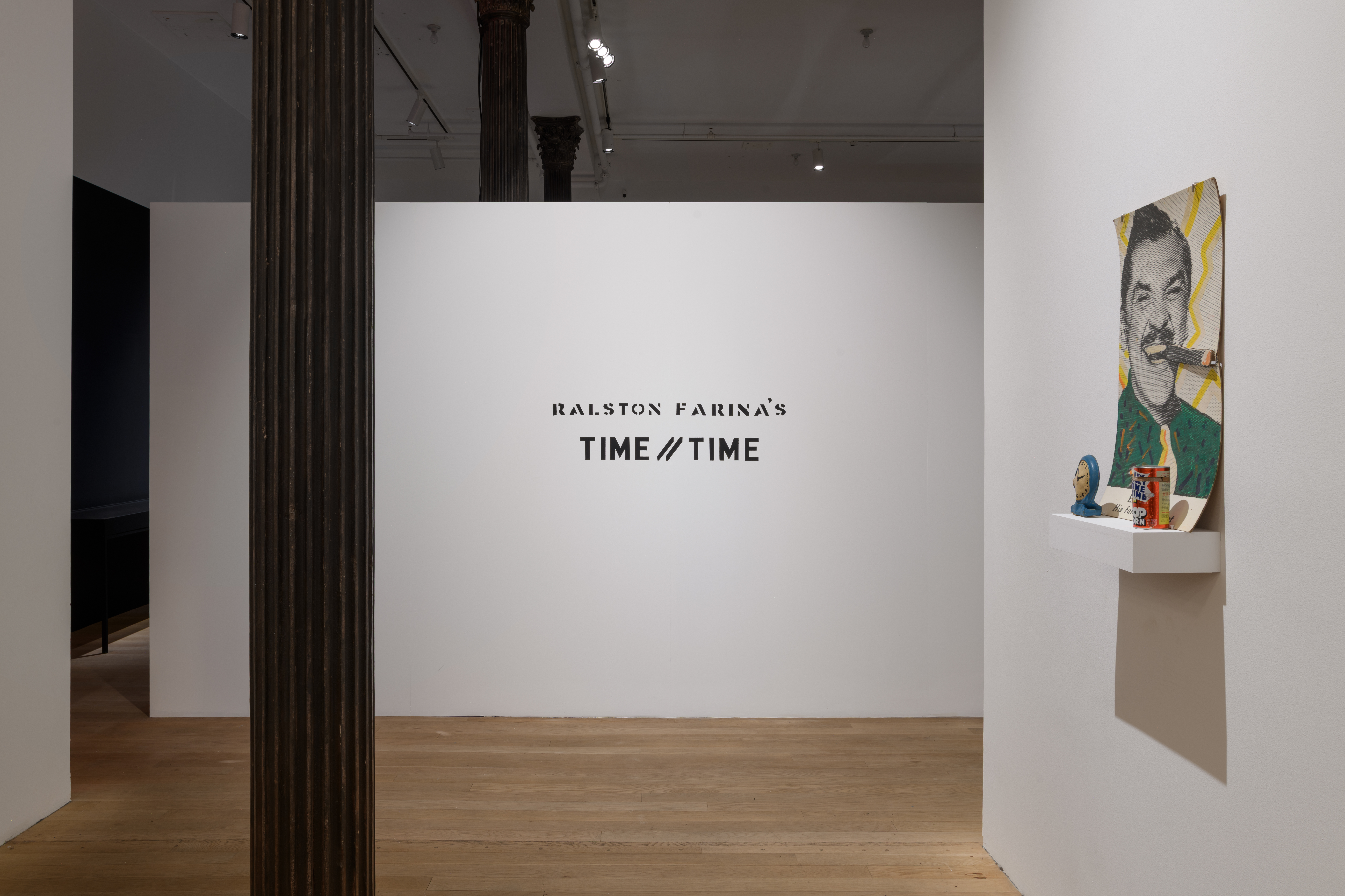 The title <em>TIME // TIME</em> is written centered on a white wall. To the right, a white shelf displays objects and a colorful poster of Ernie Kovacs.