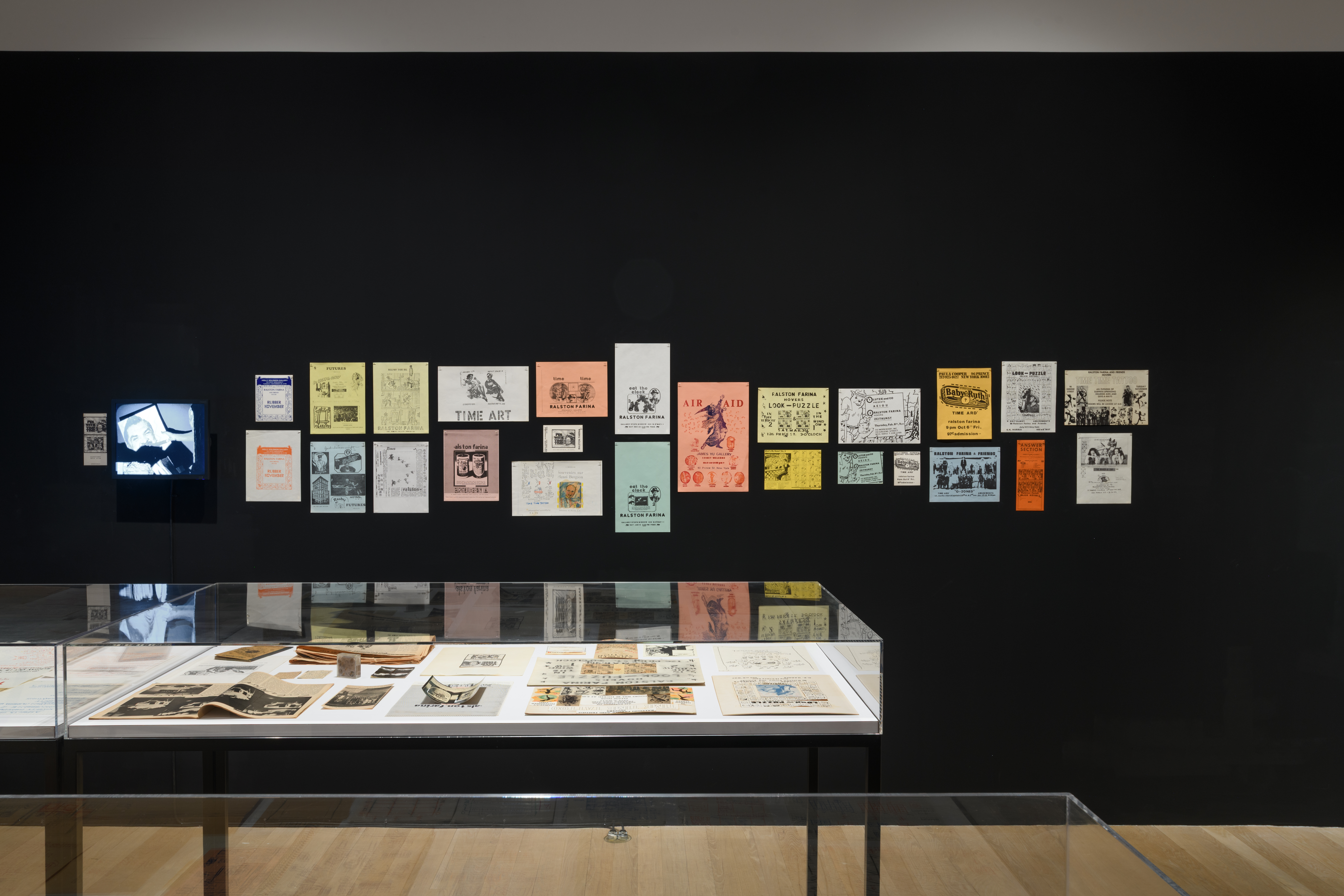 Various colorful posters hang in a disassembled row on a black wall. In front of the wall sits a large display case exhibiting newspaper clippings, flyers, and drawings.
