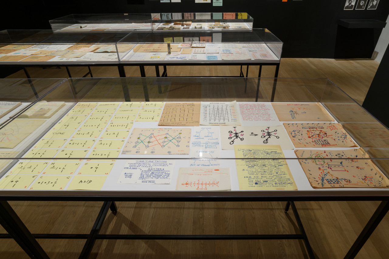 Three large display cases exhibit various objects. The case most within sight displays a selection of drawings and handwritten pages. To the left of the case, numerous yellow note cards align to form a code.