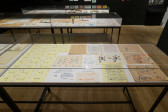 Three large display cases exhibit various objects. The case most within sight displays a selection of drawings and handwritten pages. To the left of the case, numerous yellow note cards align to form a code. 