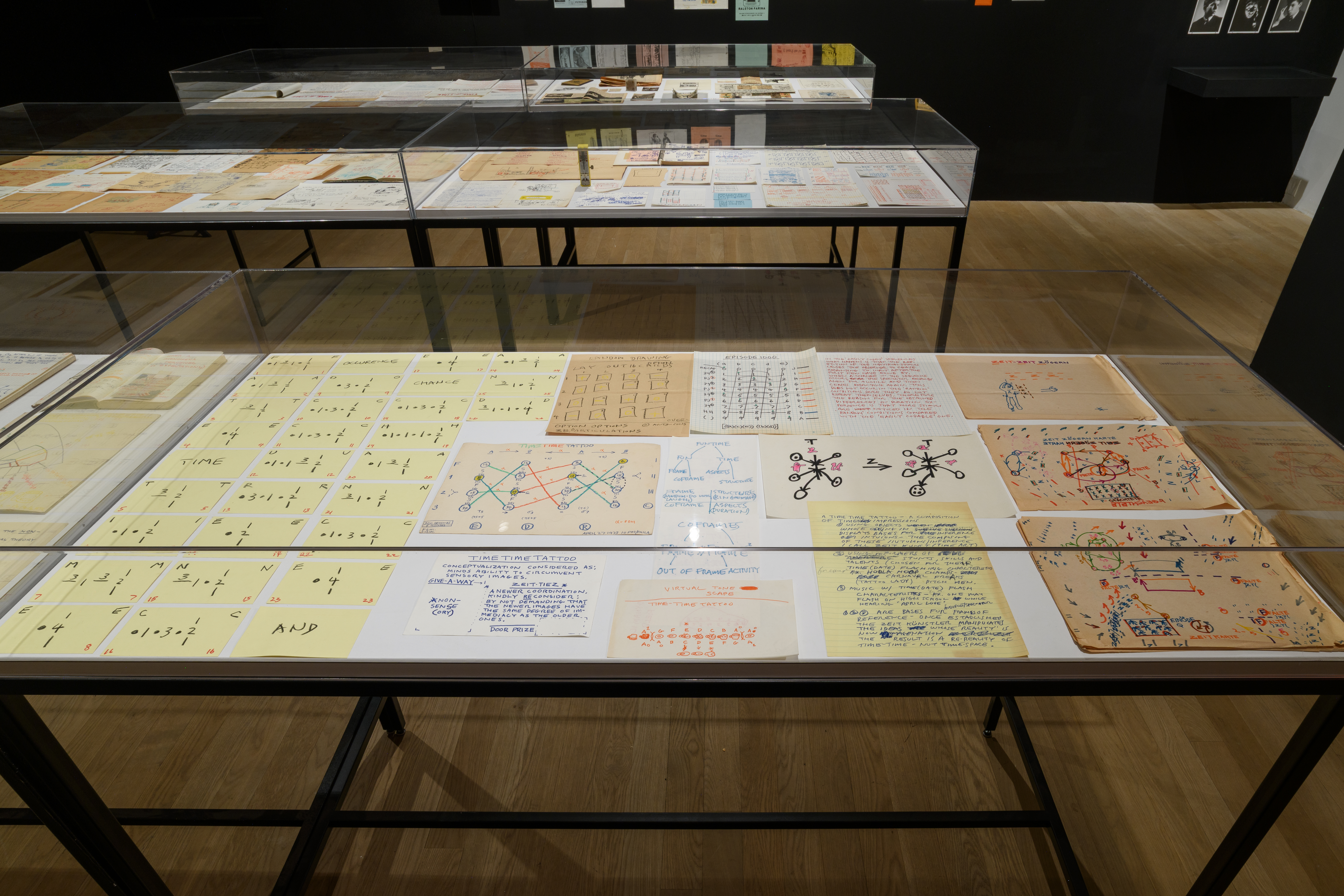 Three large display cases exhibit various objects. The case most within sight displays a selection of drawings and handwritten pages. To the left of the case, numerous yellow note cards align to form a code. 