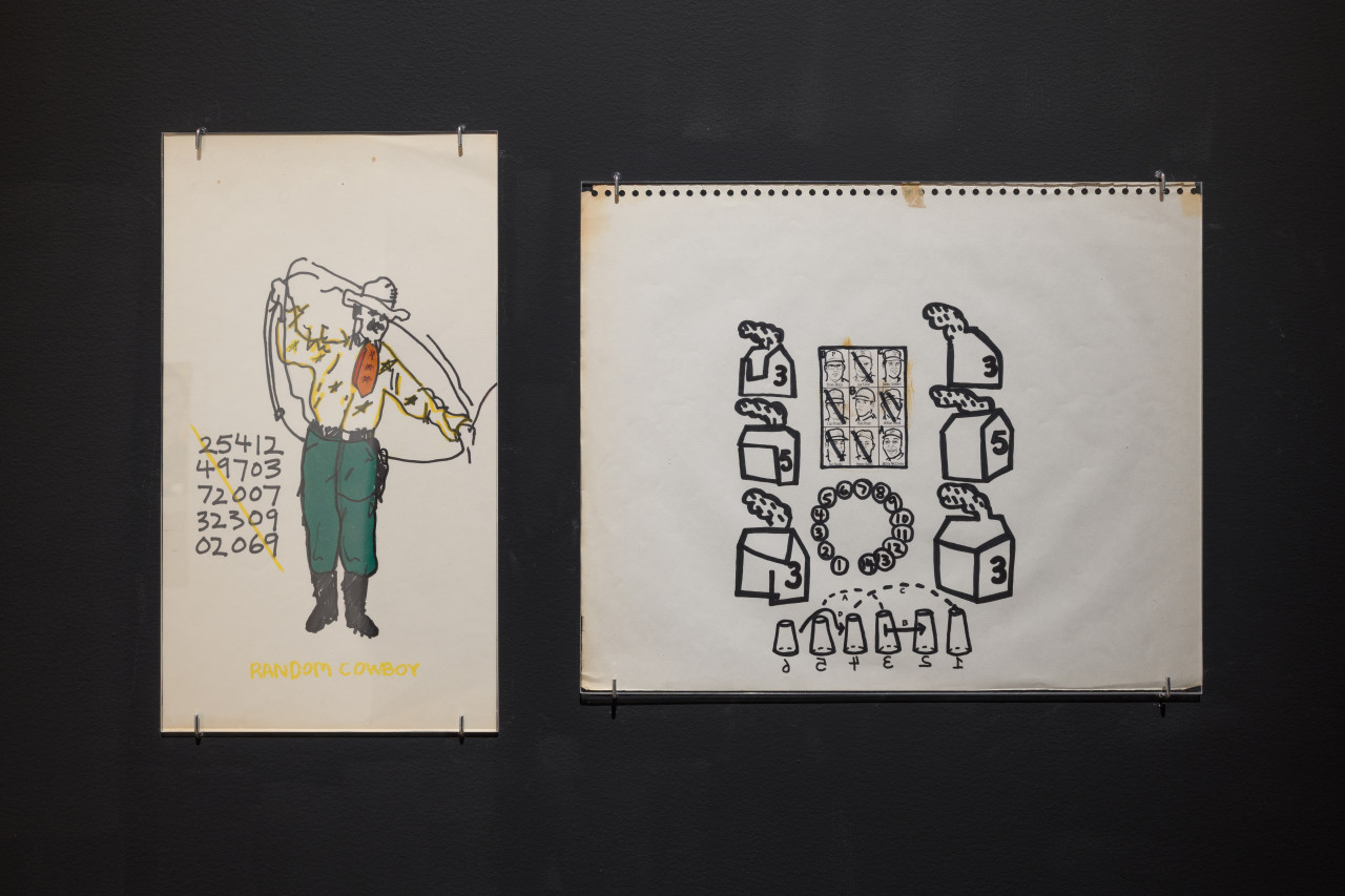 Two drawings are exhibited on a black wall. On the left, a colorful drawing of a cowboy is accompanied by a grid of numbers and titled <em>Random Cowboy</em>. On the right, abstracted numbered houses are drawn in thick black marker and surround a grid of faces.