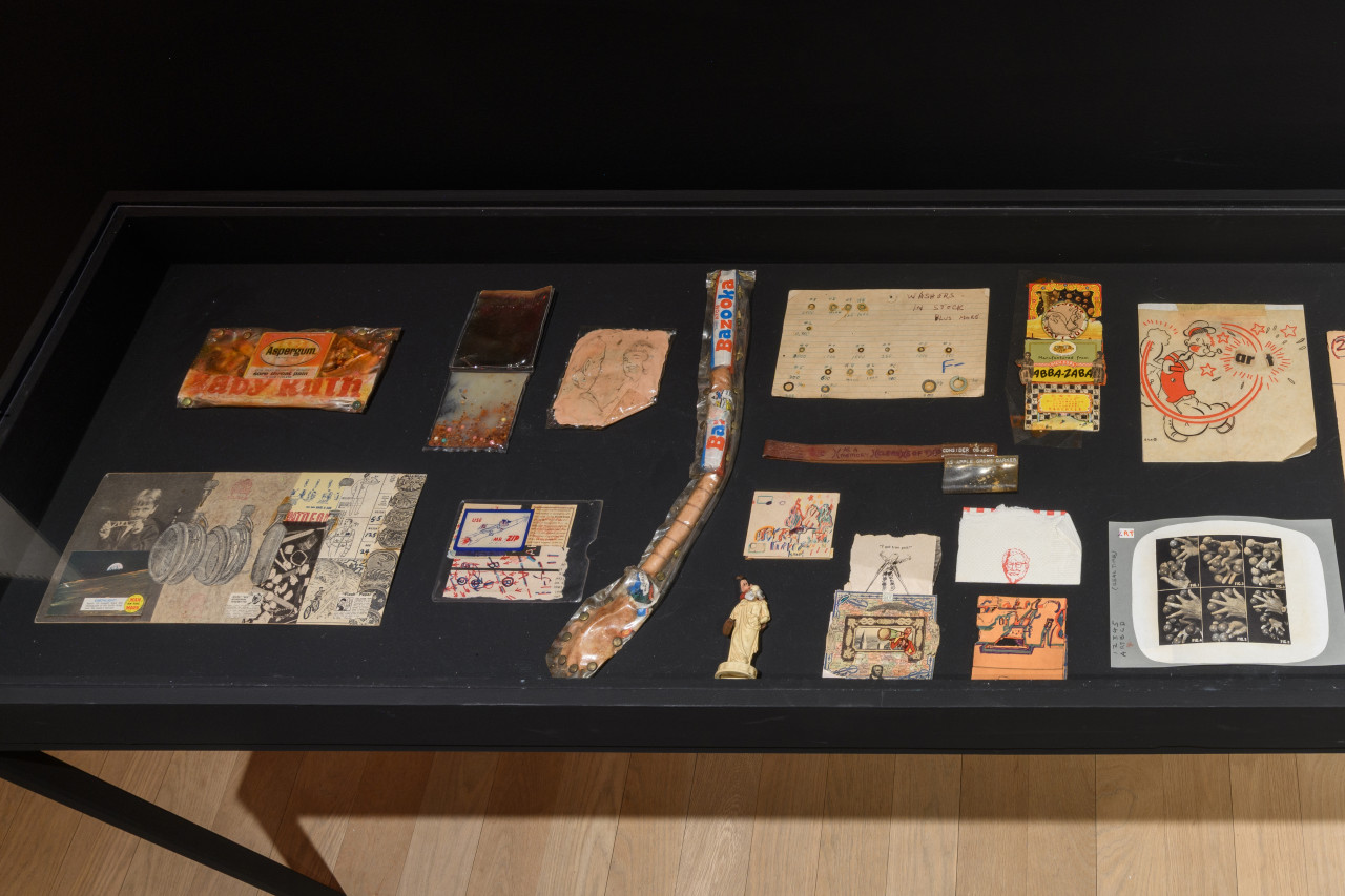A display case exhibits a variety of objects, including drawings, collages, found objects, and plastic pouches filled with various substances.