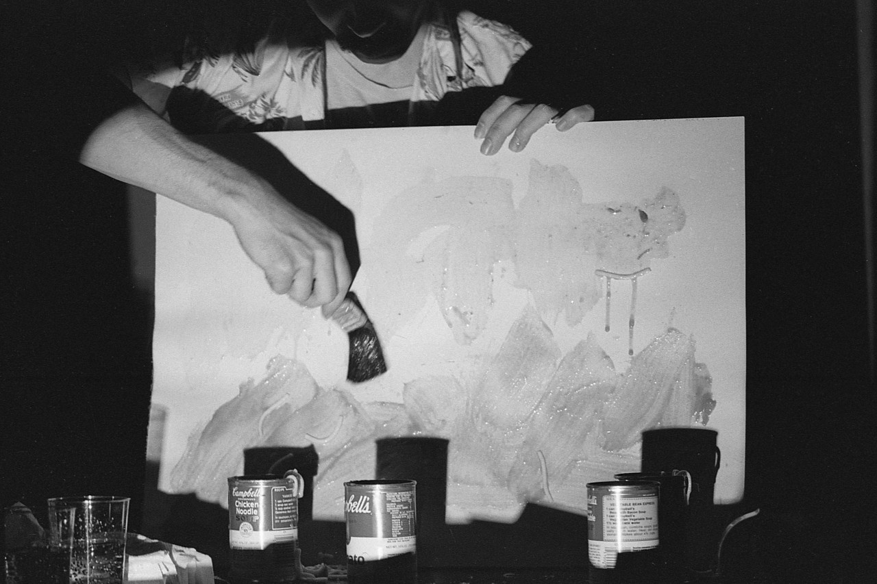 A black and white photograph of a person painting the surface of a hand-held white board with broad strokes. Sitting on the table in front are three open cans of Campbell’s Soup.