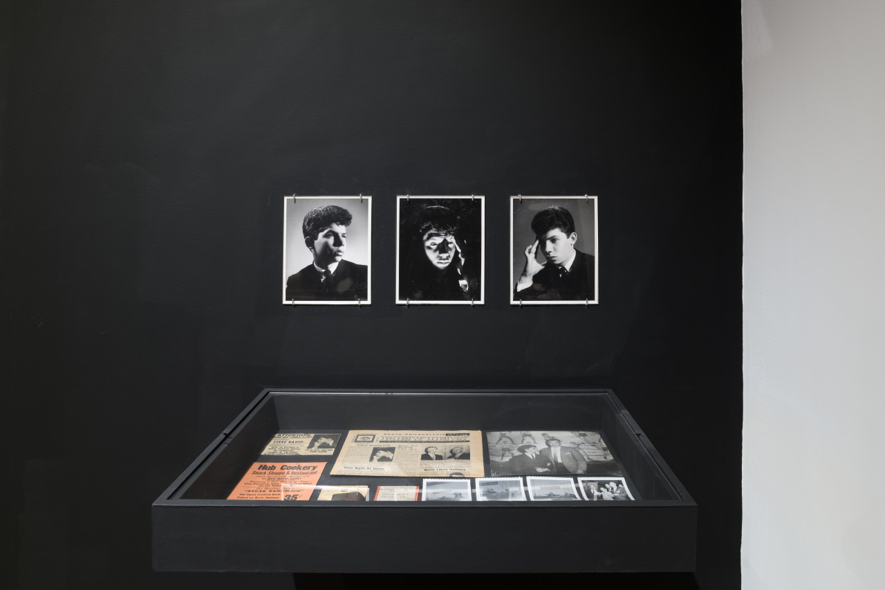 Three black-and-white photographs of a young Ralston Farina, mounted on a black wall, hang above a display case.