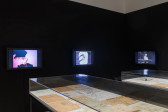 Three TV screens, mounted on a black wall, glow with various images. Each TV plays a performance by Ralston Farina. In front of the screens sit two display cases.