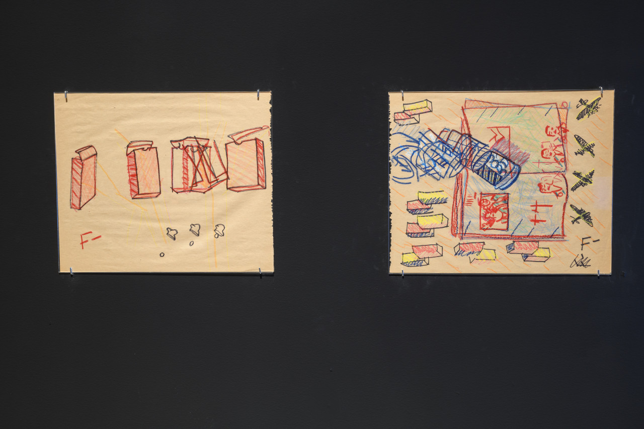 Two colorful drawings are exhibited on a black wall. On the left, a series of open boxes is drawn in red. To the right, the drawing is crowded with the depiction of an open book, airplanes, and multicolored boxes. Both drawings are signed with F-.