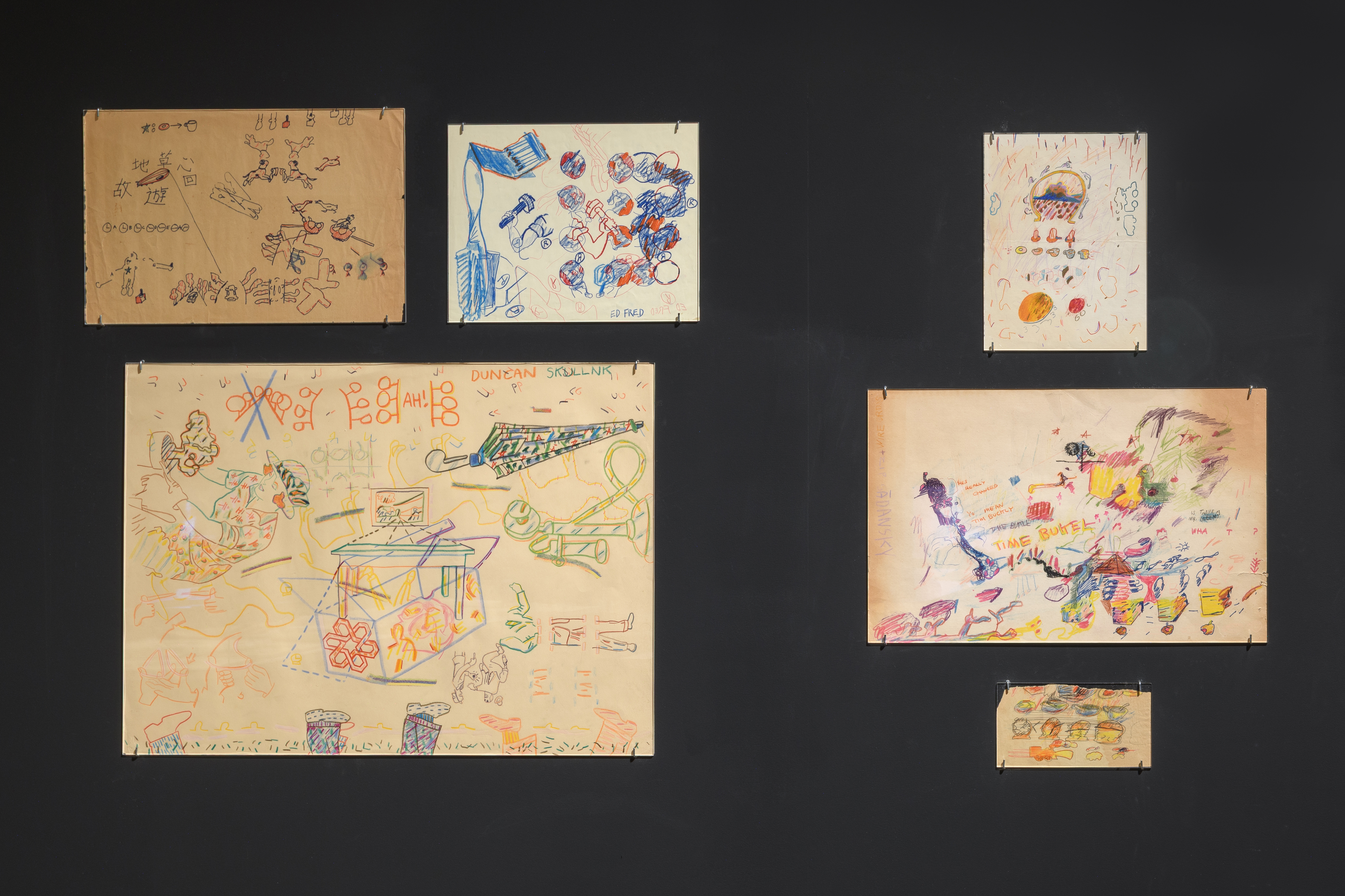 Six abstracted and colorful drawings hang on a black wall. They vary in size and include motifs of clocks, animals, and partially drawn caricature-like people. 