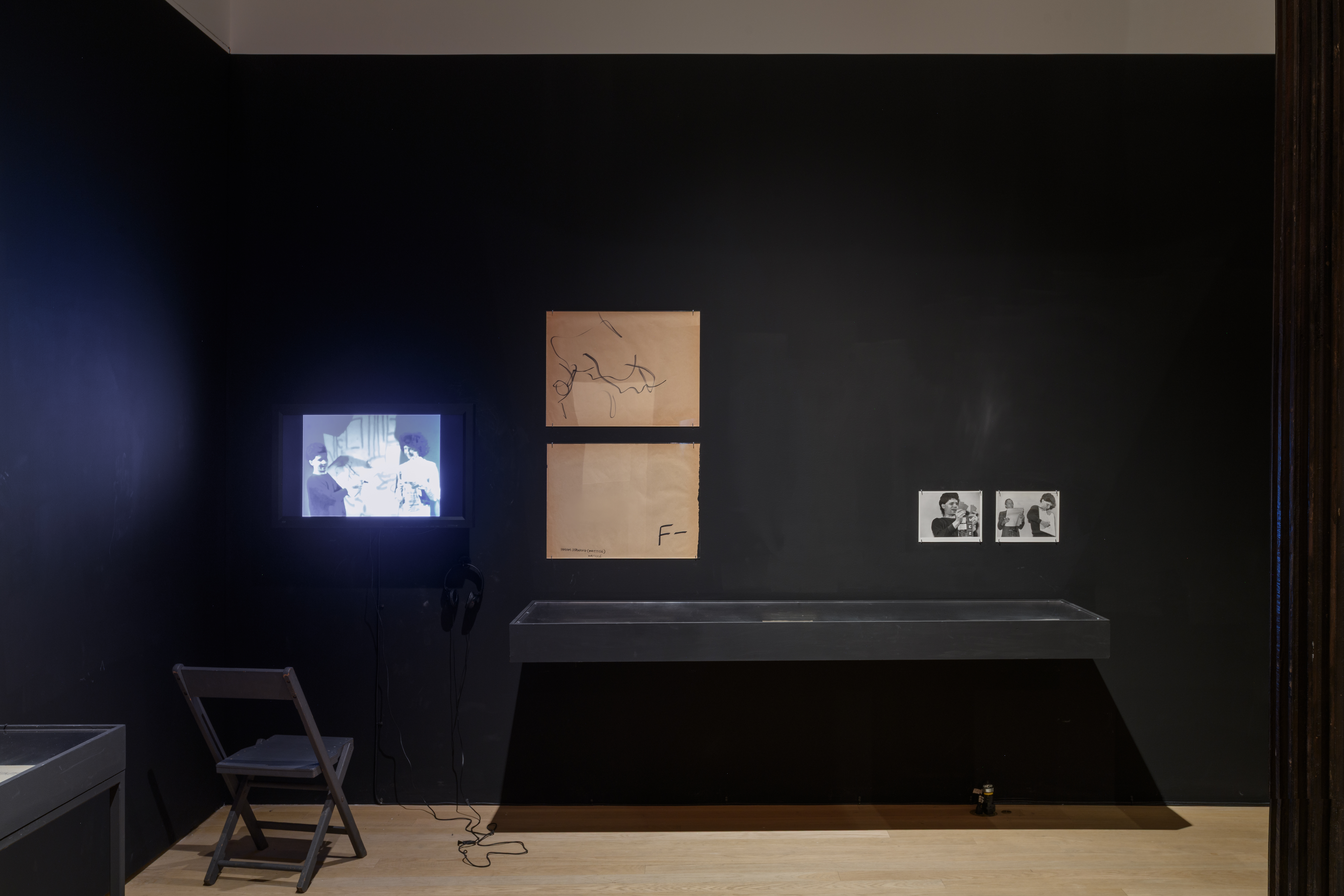 From left to right, a chair rests in front of a glowing TV screen, two larger drawings hang parallel, and two small black-and-white photographs of Ralston Farina hang beside one another.