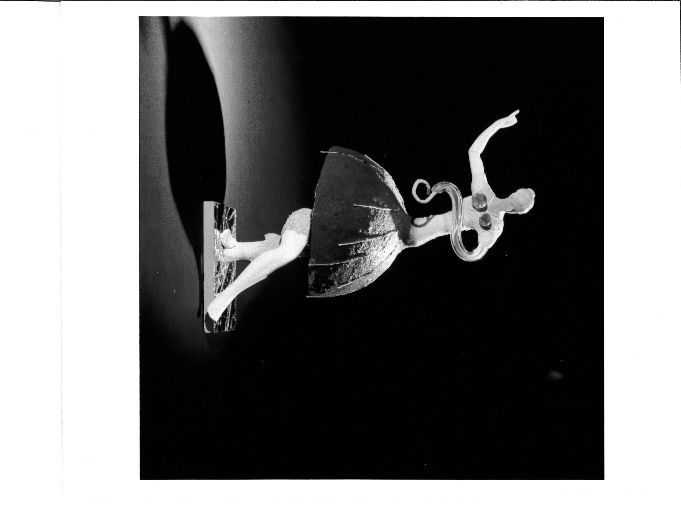 A small constructed dancing figurine appears on a pedestal with light shining on it from the right creating a shadow on its left.