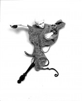 A figurine human head and hands appear from an amalgamation of various objections and materials hanging on a white wall.