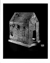 A black and white photograph of a sculpted, rough-hewn house-like object with a black background.