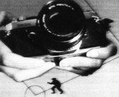 A close up image of hands holding a film camera in black and white on a white background.  Below the hands, a small, black silhouette of a figure walking.