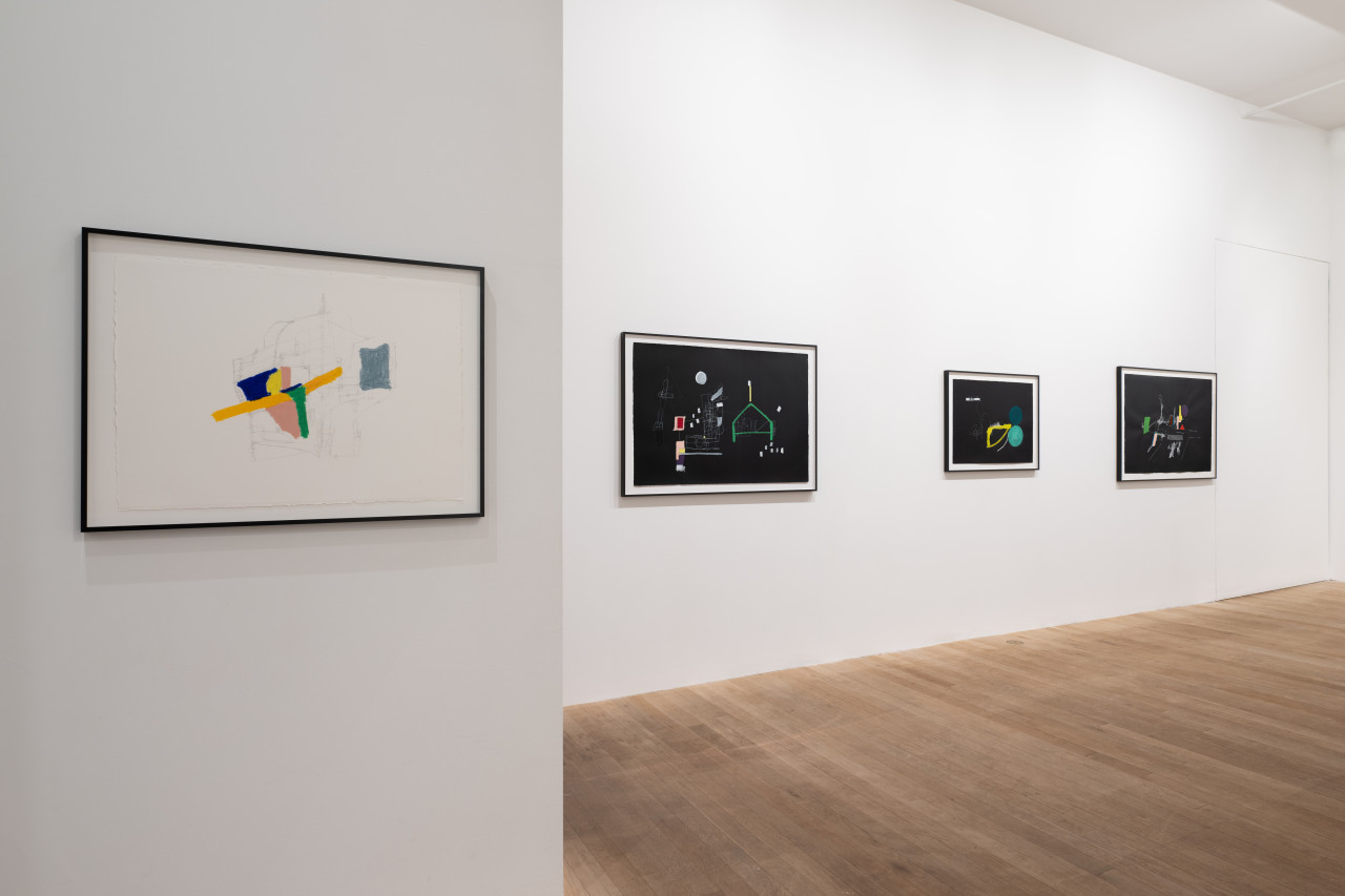 A color image of a gallery space. On the left wall hangs a framed work that consists of a colorful abstract drawing made on white paper, with pencil strokes surrounding the colored shapes. On the right wall, three framed works hang on a horizontal line. The three abstract works are made on black paper, and depict colorful compositions.