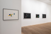 A color image of a gallery space. On the left wall hangs a framed work that consists of a colorful abstract drawing made on white paper, with pencil strokes surrounding the colored shapes. On the right wall, three framed works hang on a horizontal line. The three abstract works are made on black paper, and depict colorful compositions.