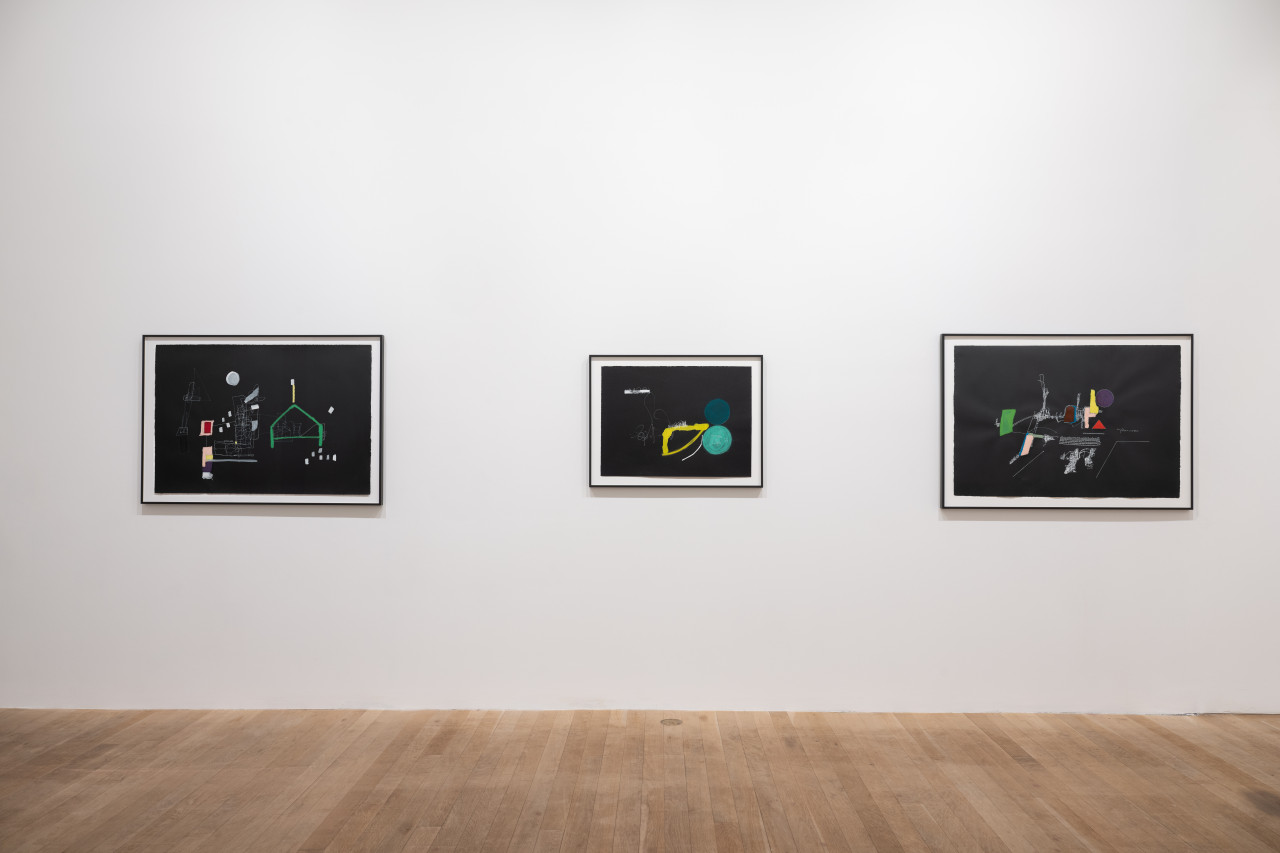 A color image of three framed works hang on the white wall. The works, all made on black paper, consist of colorful shapes, white line drawings and illegible text that form abstract compsitions.