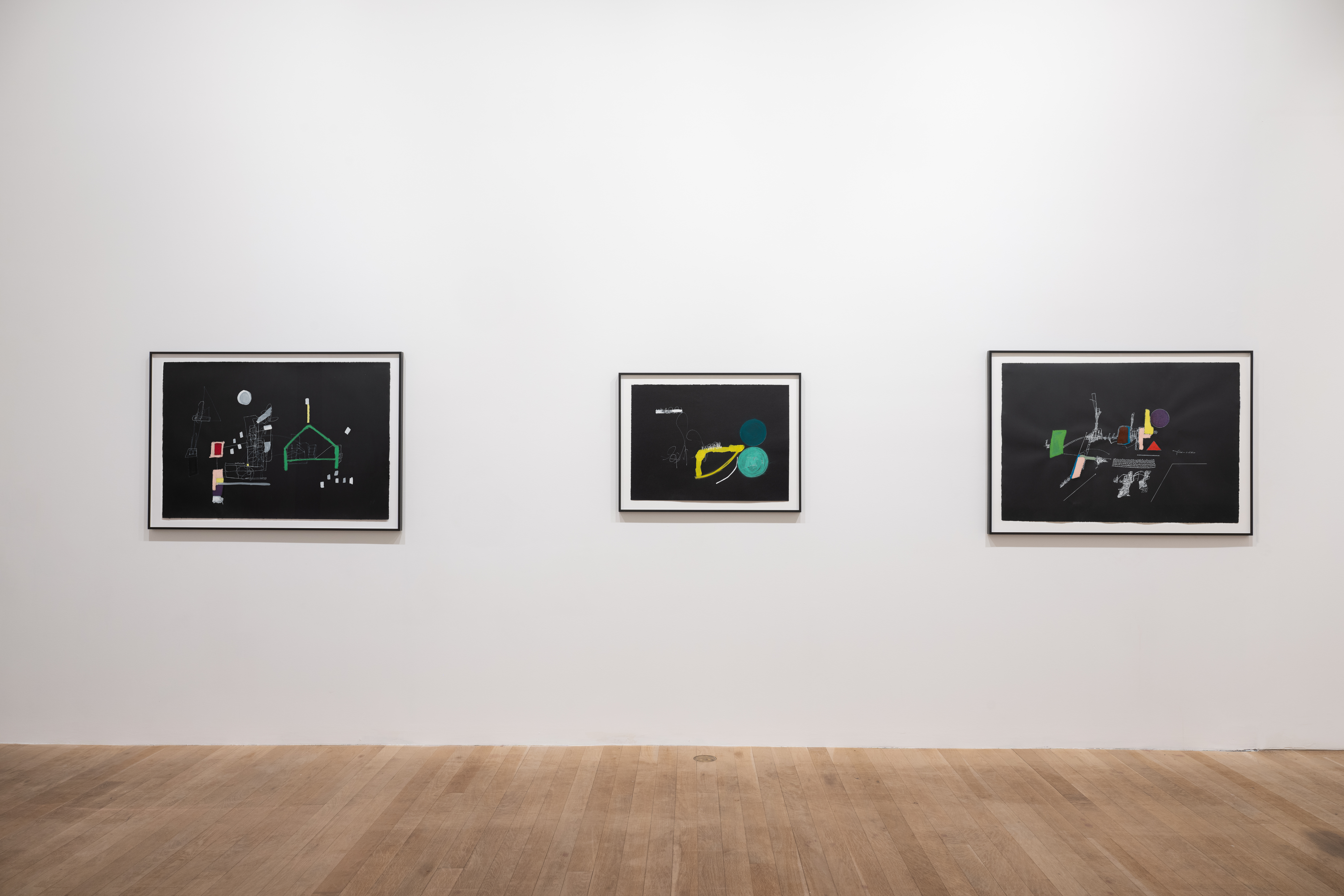 A color image of three framed works hang on the white wall. The works, all made on black paper, consist of colorful shapes, white line drawings and illegible text that form abstract compsitions.