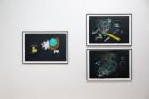 A color image of three framed works against a white wall. The drawings are done on black paper and depict abstract compositions made up of white, and colorful shapes, lines and strokes.