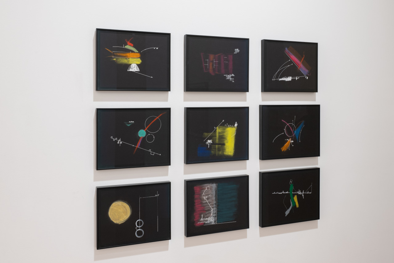 A color image of nine framed works displayed as a grid against a white wall. The drawings are done on black paper and depict abstract compositions made up of colorful shapes and strokes, and white lines and arrows.
