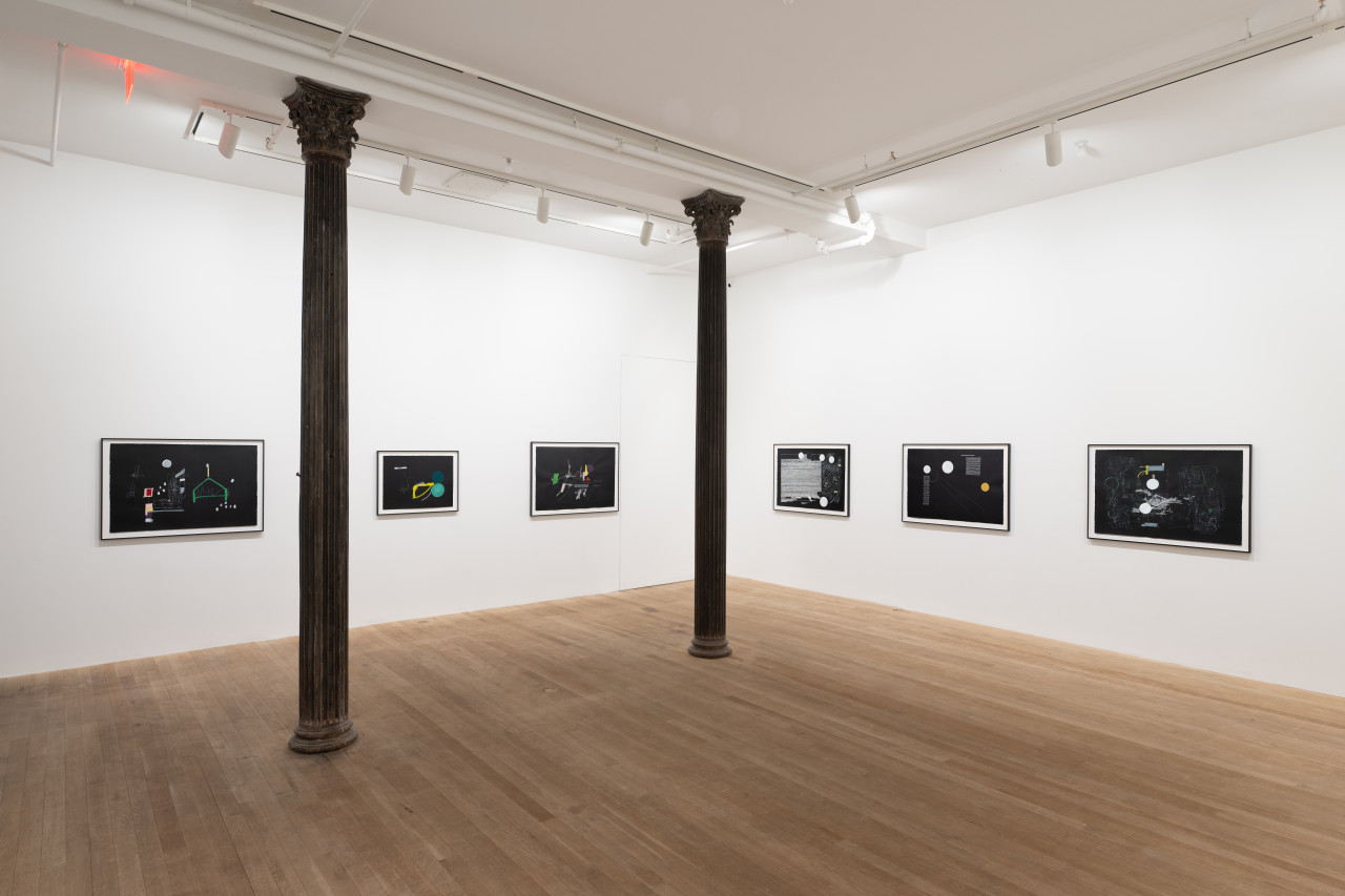 A color image of the corner of a gallery space with two columns. Across the two white walls, six framed abstract drawings on black paper are displayed in a horizontal line.