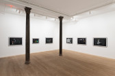 A color image of the corner of a gallery space with two columns. Across the two white walls, six framed abstract drawings on black paper are displayed in a horizontal line.